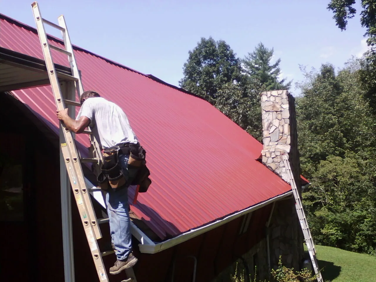 Roofer on a ladder beside a red metal roof and stone chimney — Soffit & Fascia Repair by Roofing Brothers in Greenvale